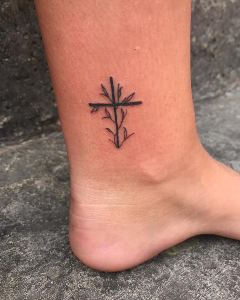 A minimalist black tattoo of a cross intertwined with delicate branches on an ankle.