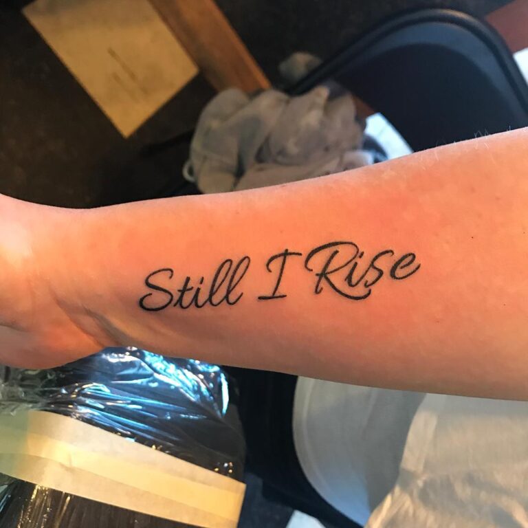 A tattoo on a forearm displays the phrase "Still I Rise" in elegant script.