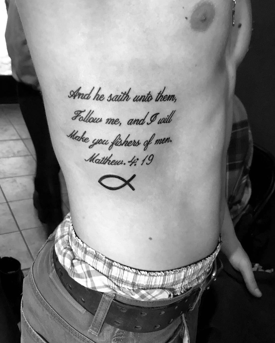 Tattoo on side featuring a biblical quote from Matthew 4:19 and a fish symbol.