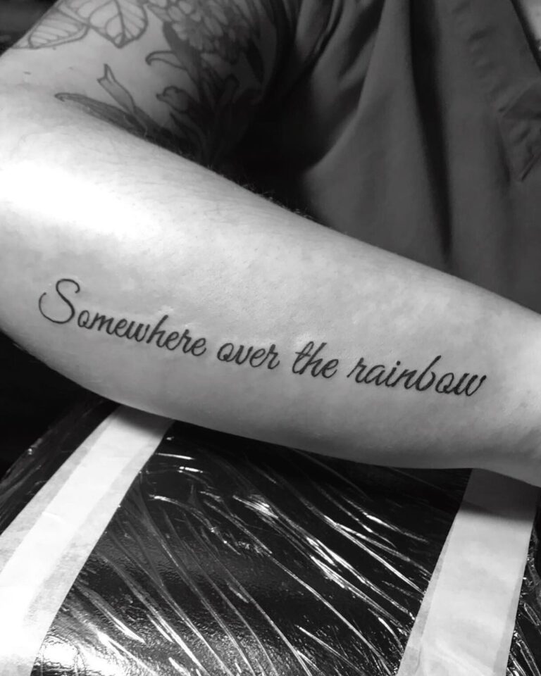 A forearm tattoo features the phrase "Somewhere over the rainbow" in elegant script.