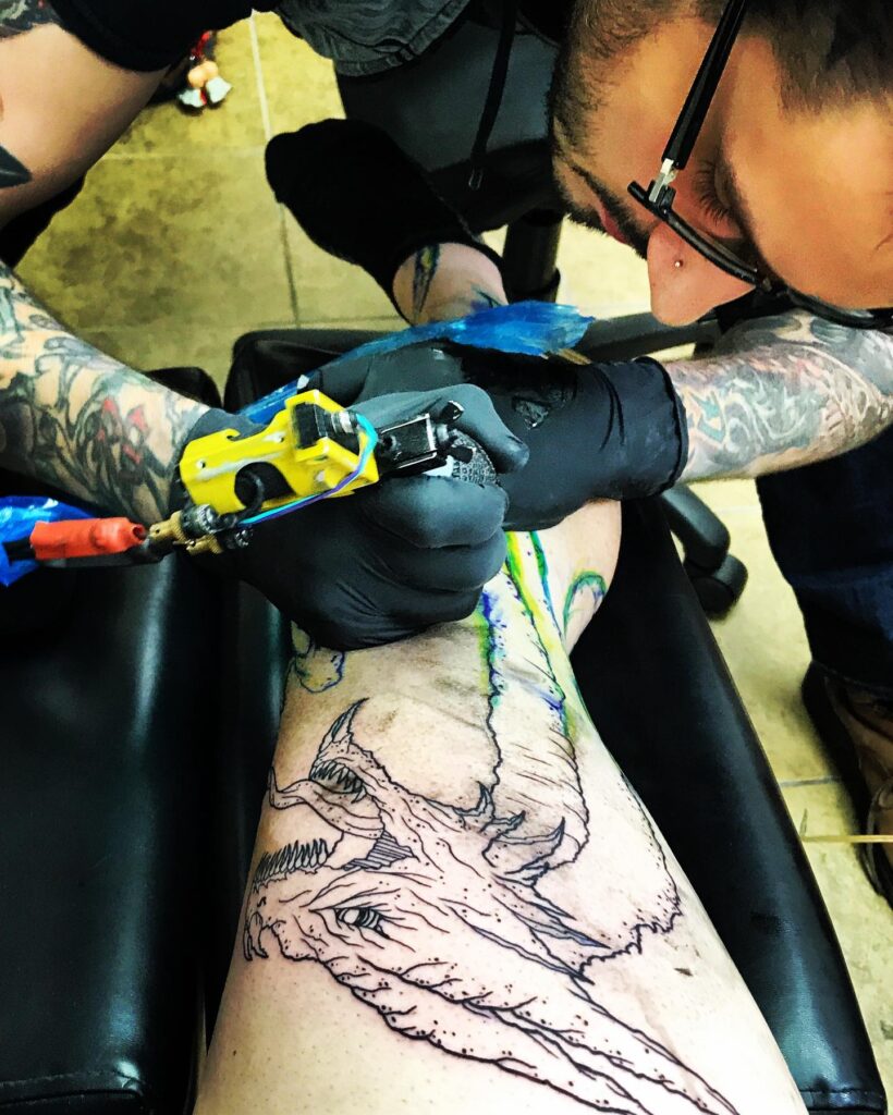 Tattoo artist inking a detailed dragon design on a client's leg, showcasing vibrant colors and intricate line work.
