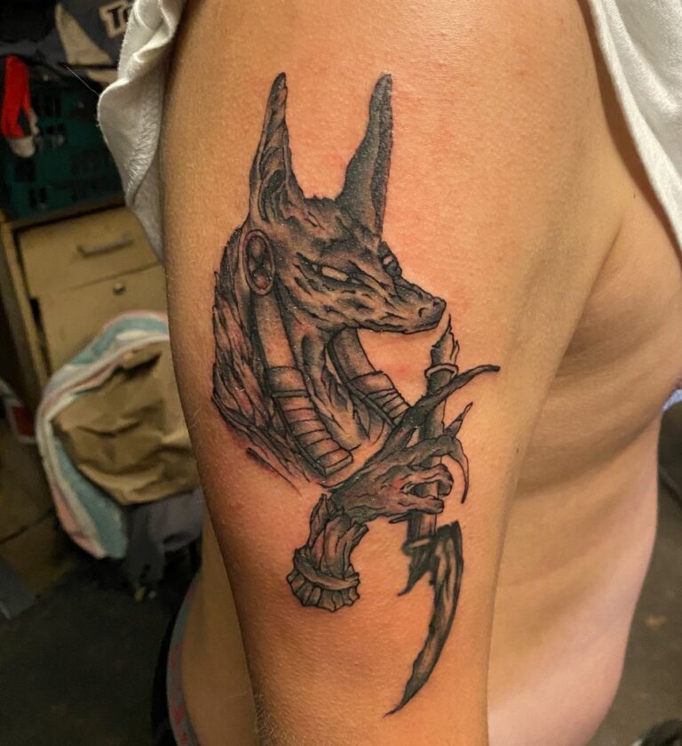 Tattoo of Anubis, the Egyptian god, holding a dagger, intricately detailed on a person's upper arm.