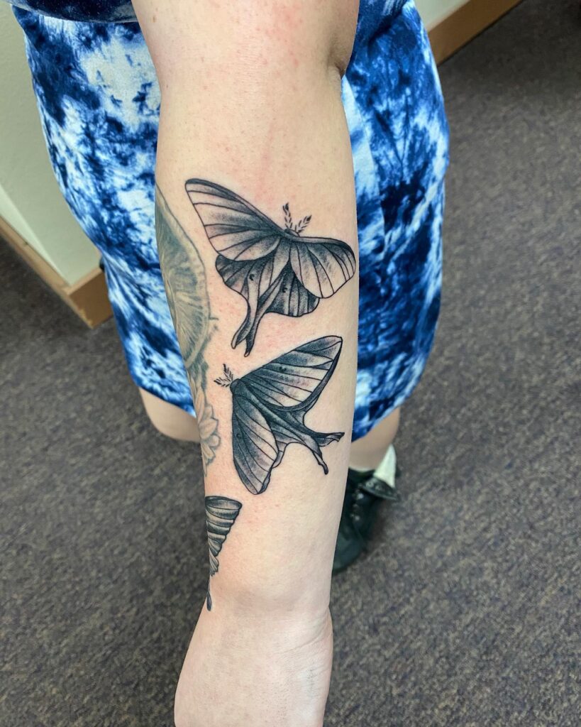 Three detailed black and gray moth tattoos adorn the forearm of a person wearing a blue tie-dye dress.