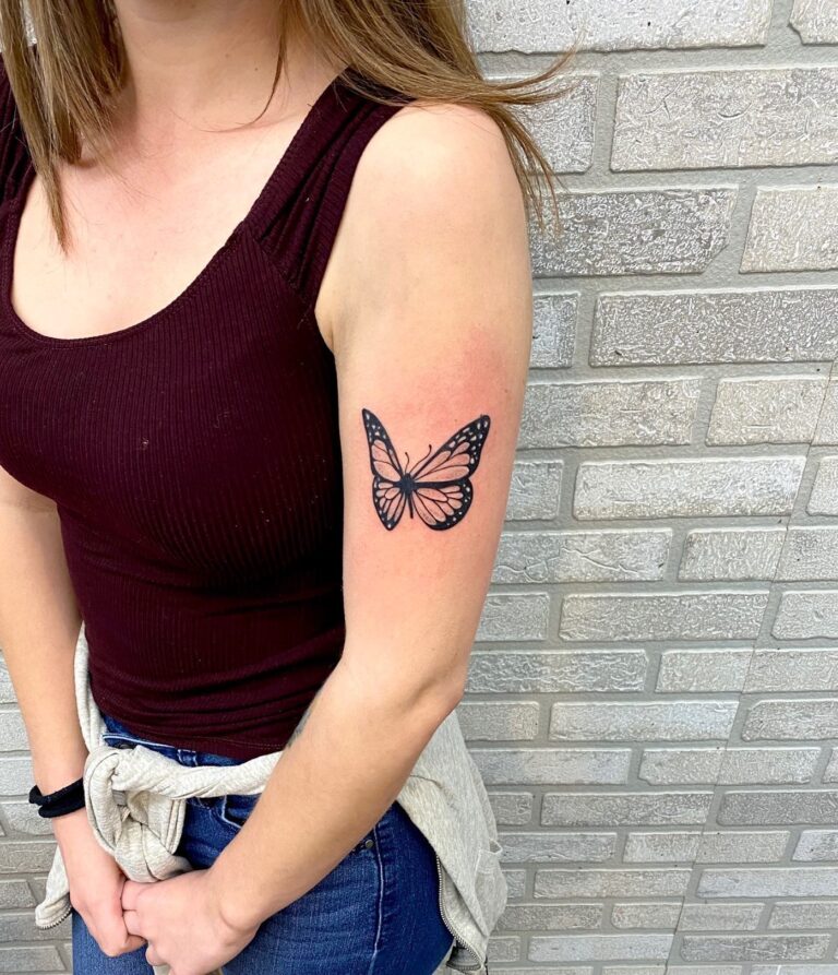 A woman shows off a butterfly tattoo on her upper arm against a textured brick wall.