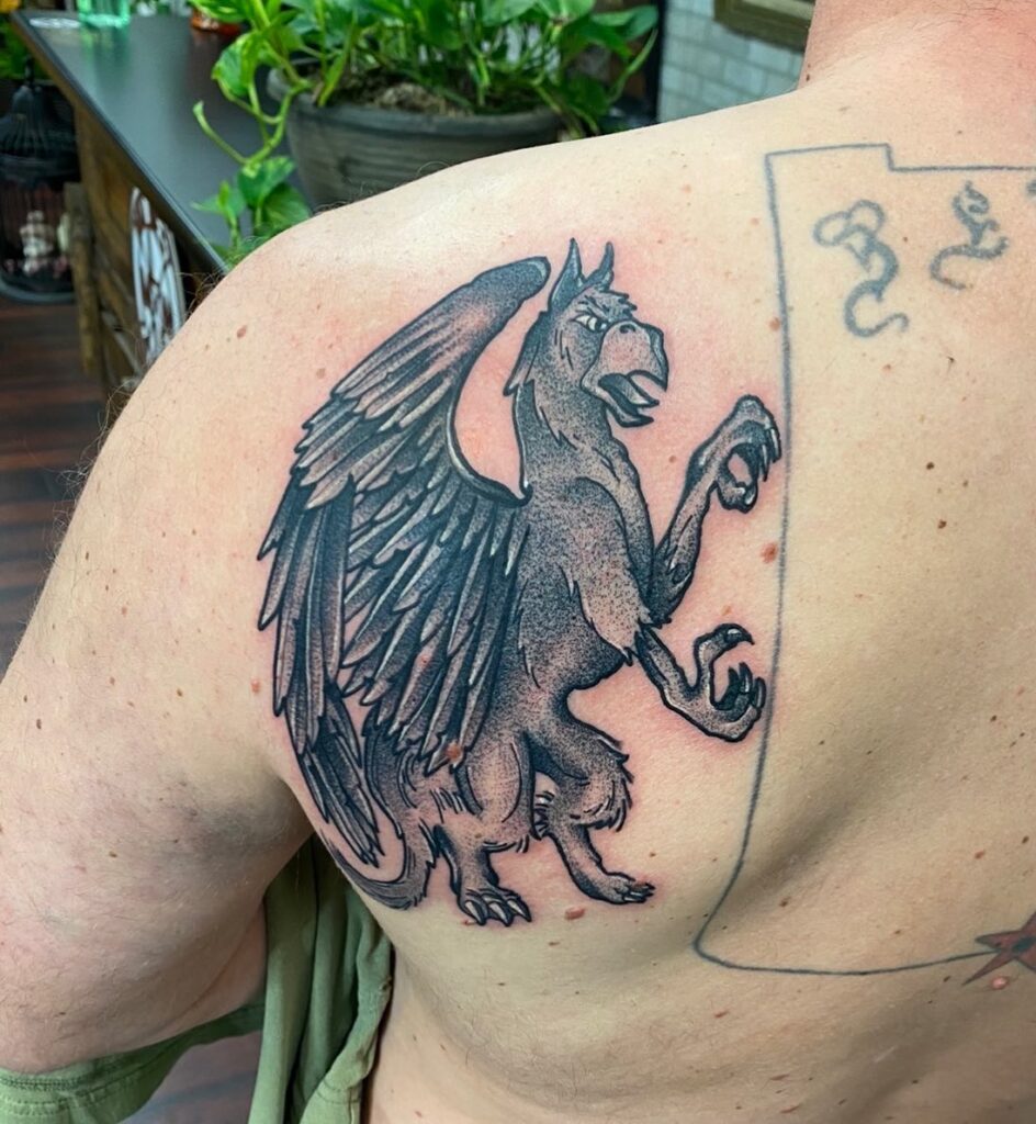 A detailed tattoo of a griffin with wings, displayed on a person's upper back in a tattoo studio setting.