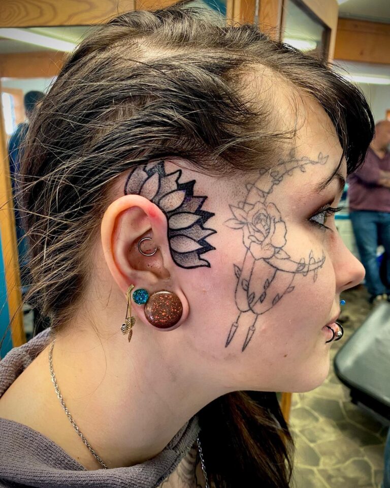 Side profile of a person with intricate tattoos, including a lotus and rose, and multiple ear piercings.