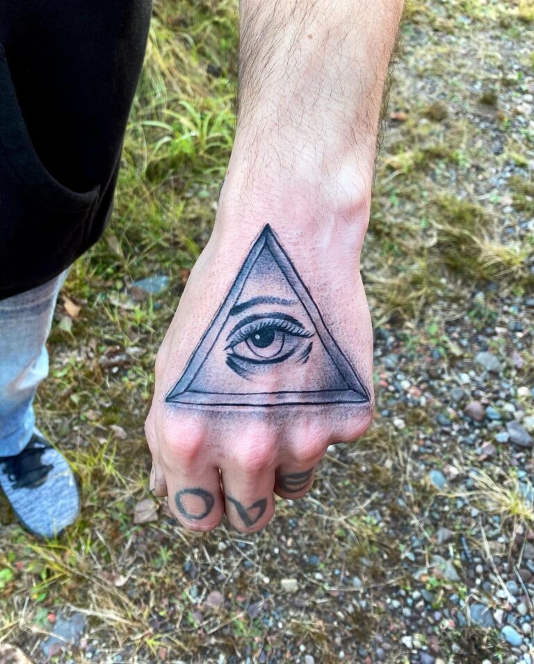 A hand with a detailed tattoo of an eye within a triangle, surrounded by grass and gravel.
