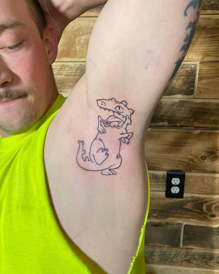 A person shows off a cartoon dinosaur tattoo on their upper arm against a wooden backdrop.