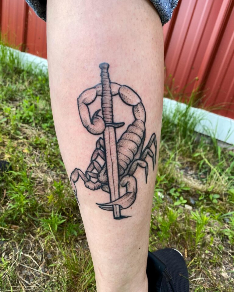 A detailed tattoo of a scorpion gripping a sword on a person's leg, set against a grassy background.