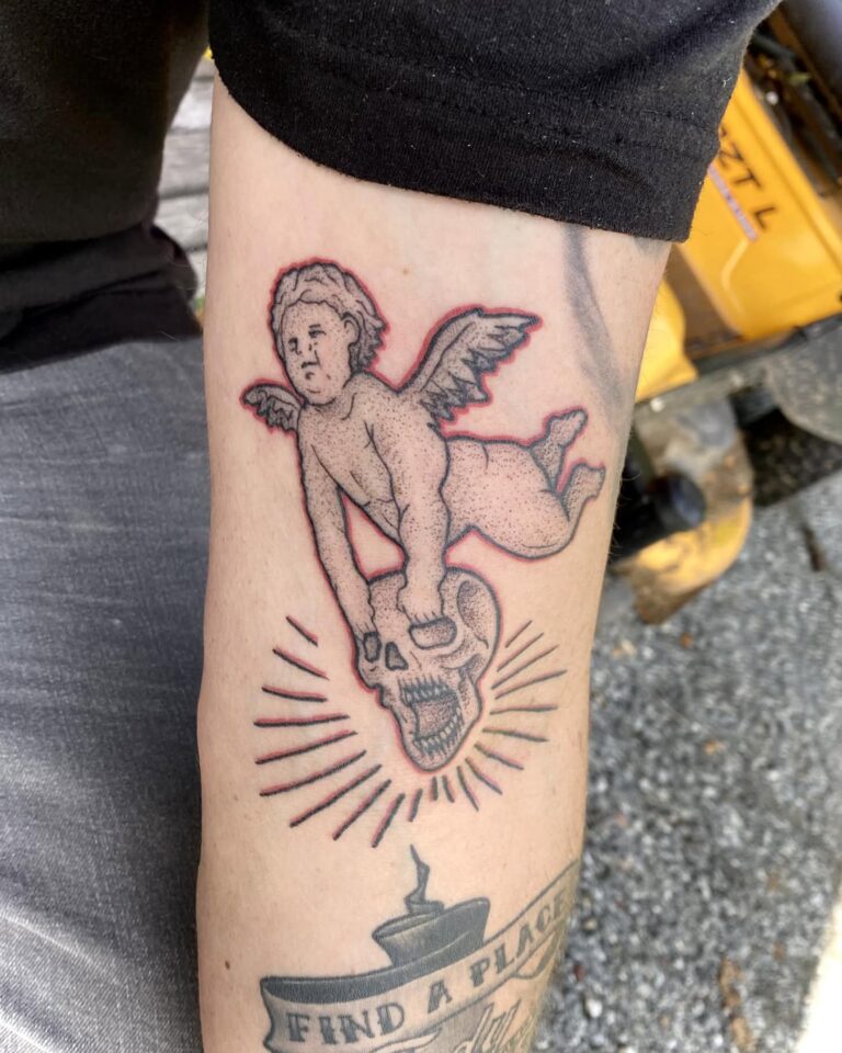 Tattoo on forearm featuring a winged cherub holding a skull, surrounded by radiating lines and text below.