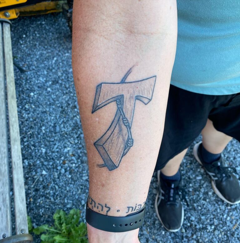 Tattoo on forearm featuring an axe intertwined with a stylized letter T and Hebrew text below.