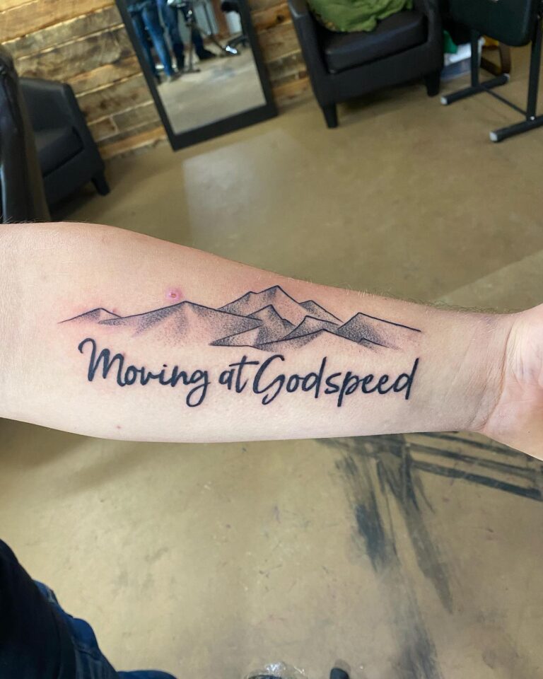 Tattoo on forearm featuring mountains and the phrase "Moving at Godspeed" in elegant script.