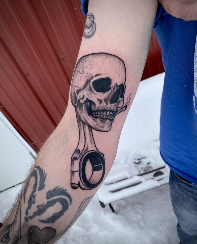 A detailed tattoo of a skull with a cigarette, integrated with a mechanical piston design on a person's forearm.