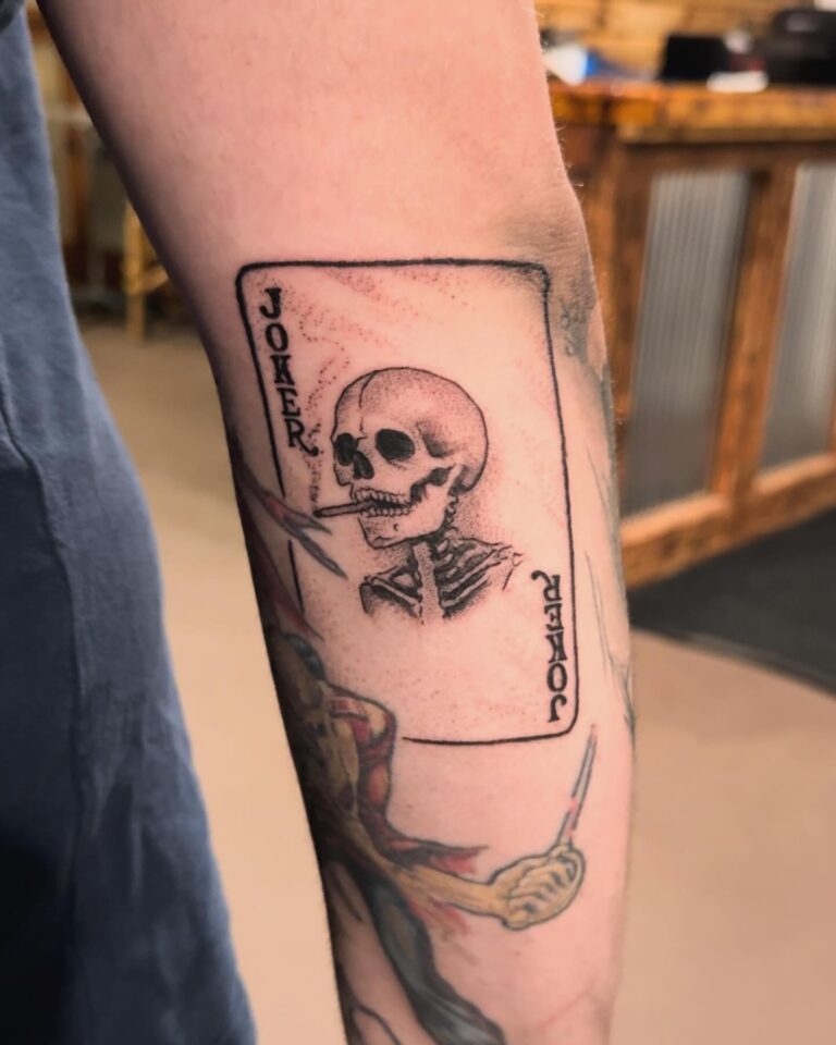 Tattoo of a skeleton on a Joker playing card, featuring intricate details and a smoking cigarette.