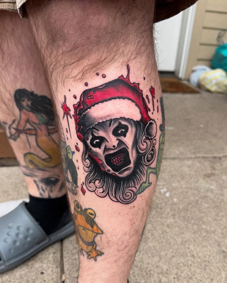 A leg tattoo features a menacing face wearing a Santa hat, surrounded by splashes of red ink and other colorful designs.
