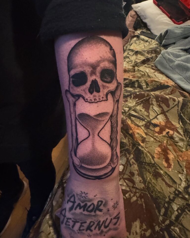 A detailed tattoo featuring a skull and hourglass with the phrase "Amor Aeternus" on the forearm.