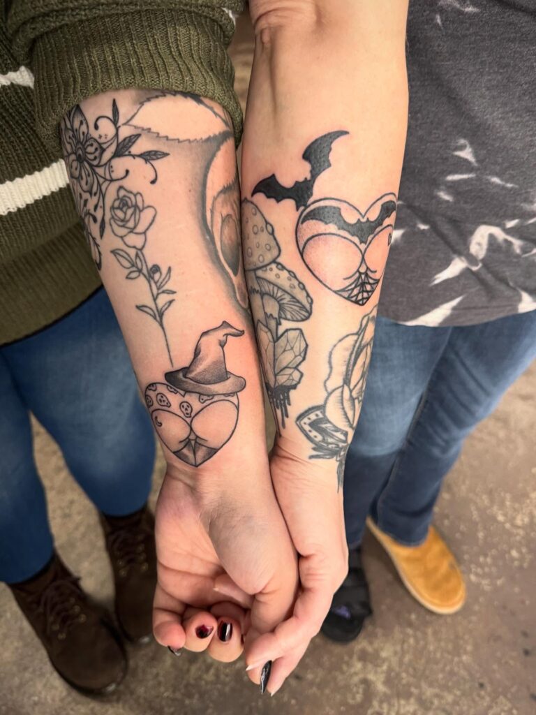 Two people hold hands, showcasing intricate tattoos of hearts, flowers, and a bat on their forearms.