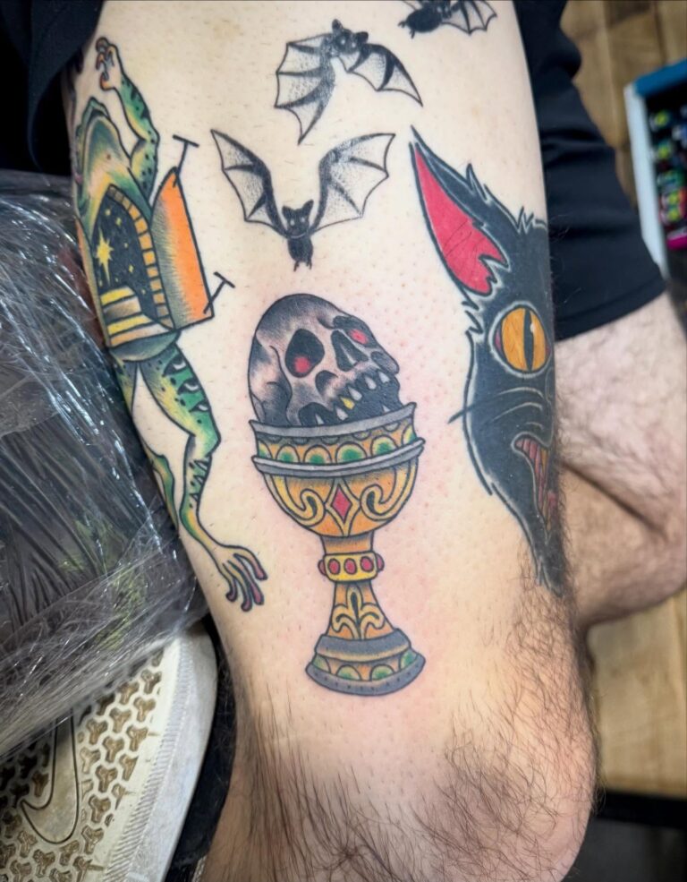 Colorful tattoos featuring a skull in a chalice, bats, a frog, and a black cat on a person's leg.