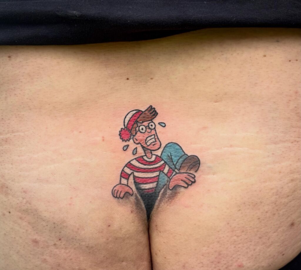 Tattoo of a cartoon character resembling Waldo, appearing to emerge from a person's lower back.